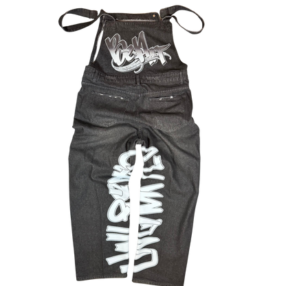 Front view of black distressed BeArt overalls featuring ripped denim, graffiti lettering down the leg, and adjustable straps.