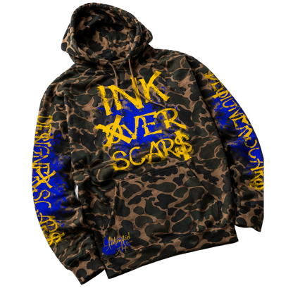 A camouflage hoodie with a brown and black animal print pattern, featuring bold yellow text reading 'INK AVER SCARS' on the front and blue text down the sleeves. The brand 'Inkscars' is visible in a stylized signature near the bottom pocket.