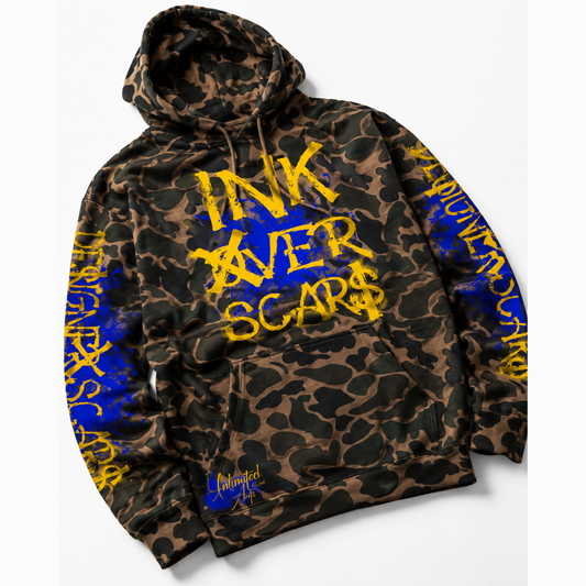 A camouflage hoodie with a brown and black animal print pattern, featuring bold yellow text reading 'INK AVER SCARS' on the front and blue text down the sleeves. The brand 'Inkscars' is visible in a stylized signature near the bottom pocket.